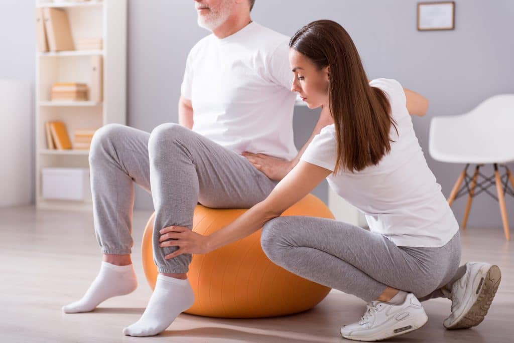 Rehabilitation. Cropped picture of male patient sitting on gym ball with concentrated female physiotherapist performing some rehab exercises